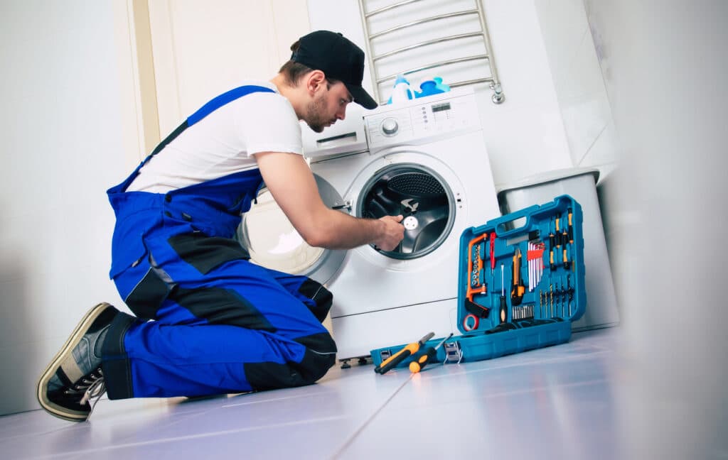 The young handsome repairman in worker suit with the professional tools box is fixing the washing machine in the bathroom